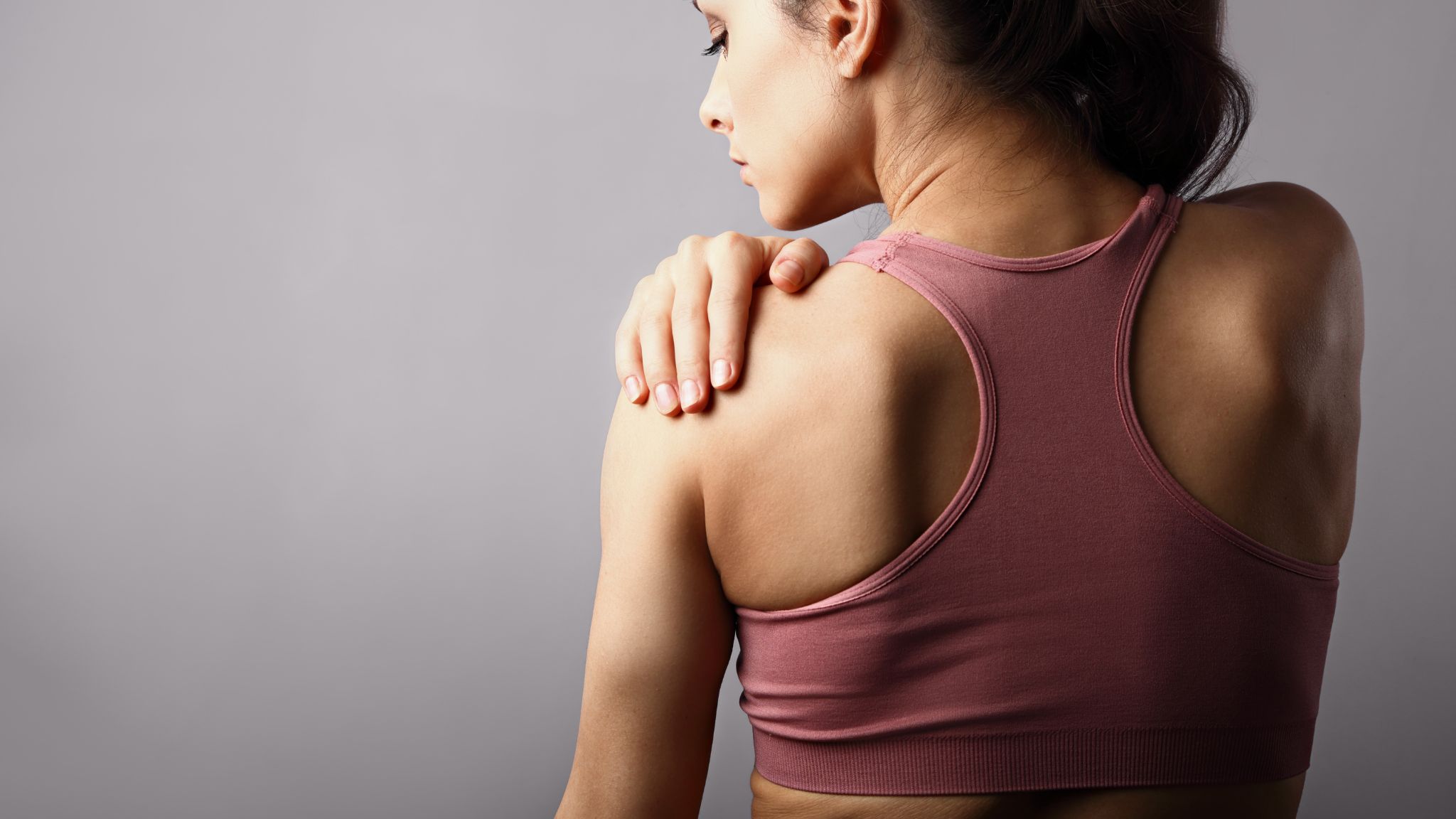 Young strong sporty woman suffering from pain in shoulder in sport wear. Touching and massaging the hand. Sports exercising injury. Closeup portrait on purple background with empty copy space 